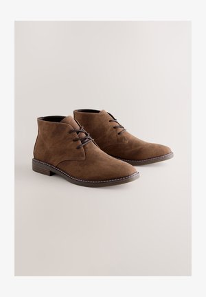 Botines de ante marrón con punta redondeada, dos cordones y tacón bajo. Presentan costuras contrastantes a lo largo de la suela. Forro interior liso.