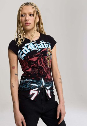 Młoda osoba z długimi blond warkoczami, ubrana w czarny T-shirt z czerwono-niebieskim wzorem i czarne spodnie, stojąca na jednolitym tle.