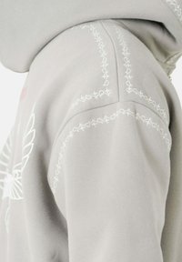 Grijze hoodie met witte geborduurde accenten langs de naden, voorzien van een subtiel ontwerp op de voorkant en een zachte, getextureerde stof.