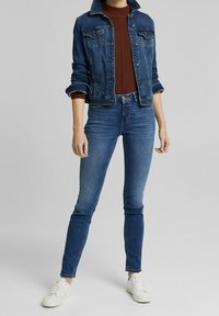 Chaqueta vaquera de color azul oscuro con dos bolsillos en el pecho, combinada con un suéter color óxido y jeans ajustados de color azul claro. Zapatillas blancas completan el conjunto.