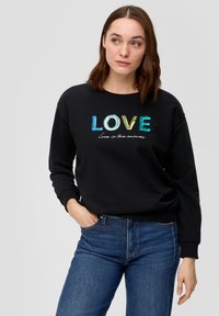 Felpa nera con grafica "LOVE" multicolore e testo bianco, con polsini aderenti, scollatura rotonda, indossata con jeans blu.