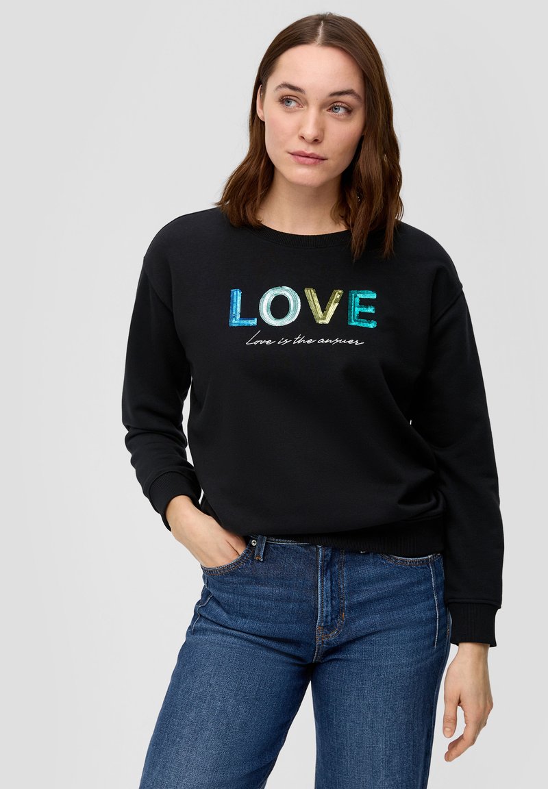 Felpa nera con grafica "LOVE" multicolore e testo bianco, con polsini aderenti, scollatura rotonda, indossata con jeans blu.