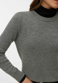 Sivi pamučni džemper s okruglim izrezom i rebrastim manšetama, slojevit preko crnog rolka. Mekana tekstura, uski kroj i minimalni detalji.