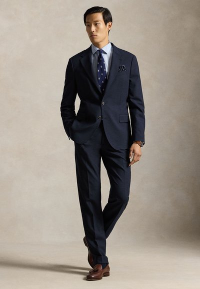 Κοστούμι σε ναυτικό μπλε με εφαρμοστή γραμμή, διαθέτει γραβάτα με εγκοπή, κλείσιμο με ένα κουμπί και ταιριαστό παντελόνι. Διακοσμημένο με γραβατάκι με σχέδιο και μαντηλάκι τσέπης.