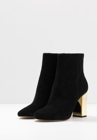 Botas de tobillo de ante negro con punta afilada y un chunky tacón metálico dorado. Textura suave sin acentos ni patrones visibles.