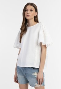 Hvid tekstureret top med bølgede ærmer og kant, parret med slidte denimshorts. Toppen har en løs, afslappet pasform.