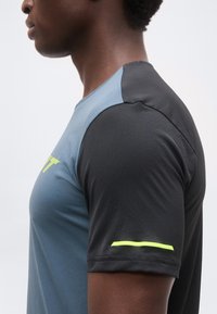 Προφίλ ατόμου που φοράει αθλητική μπλούζα με κοντά μανίκια, με ανοιχτό μπλε εμπρός μέρος και σκουρόγριμα μανίκια, διακοσμημένη με μια μικρή κίτρινη ρίγα.