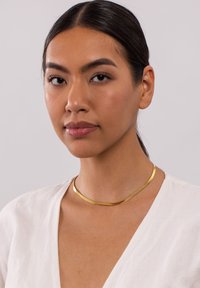 Goldfarbener Metall-Choker-Halskette mit einem glatten, geschwungenen Design. Die Oberfläche ist poliert mit einer subtilen Textur und wird über einem weißen Oberteil getragen.