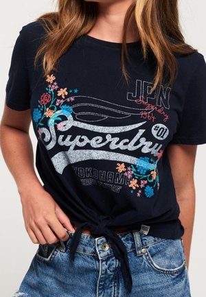 Kobieta nosząca granatowy t-shirt Superdry z kwiatowymi wzorami, związany w węzeł w talii, w połączeniu z jasnoniebieskimi jeansami.