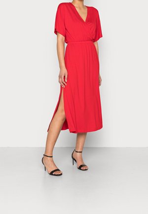 Donna che indossa un vestito midi rosso con maniche corte, scollo a V, spacco laterale e sandali neri con tacco alto e cinturino alla caviglia, in piedi su sfondo bianco.