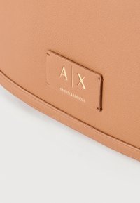 Τσάντα από δερμάτινη υφή ταμπά με ραμμένο έμπλαστρο που φέρει το χρυσό λογότυπο και τη σήμανση Armani Exchange σε λευκή επιφάνεια.