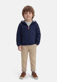 Νέος αγόρι που στέκεται φορώντας μια μπλε σκούρη ζακέτα με κουκούλα, γκρι πουκάμισο, μπεζ παντελόνι, κάλτσες με σχέδιο και καφέ αθλητικά σε λευκό φόντο.