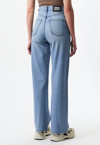 Vaaleansiniset korkeavyötäröiset farkut, joissa on suora lahje, hento haalistuminen, kaksi takataskua ja metalliset yksityiskohdat, yhdistettynä harmaisiin lenkkareihin.