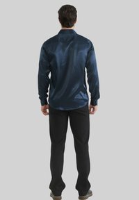 Chemise longue en satin bleu marine à manches longues avec des poignets à bouton, ourlet incurvé et texture lisse, associée à un pantalon noir au design ajusté.