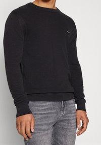 Maglione lungo nero a maniche lunghe con scollo a girocollo e polsini a coste. Piccolo logo sul petto, abbinato a jeans grigi.