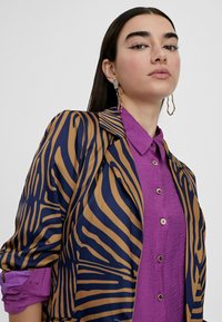 Blazer with navy and gold striped pattern, combined with a textured purple button-up shirt. Features long sleeves and large earrings.