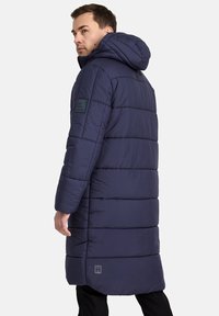 Giacca puffer di un blu navy scuro con cappuccio richiudibile con zip, design trapuntato, lunghezza lunga e cuciture rettangolari. Presenta un patch del logo sulla manica.