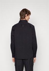 Camisa negra de manga larga con un dobladillo recto, cuello puntiagudo, puños con botón y un pequeño detalle de logo en la parte posterior del cuello.