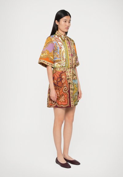 Femeie îmbrăcată cu o rochie cămașă cu model paisley multicolor, având un cordon în talie și pantofi de culoare burgundă, stând în fața unui fundal alb.