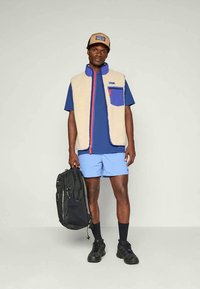 Beige boleró, kék kiemelésekkel, cipzáras zsebbel, kék pólóval és rövidnadrággal, fekete hátizsákkal, fekete sportcipővel és bézs sapkával.