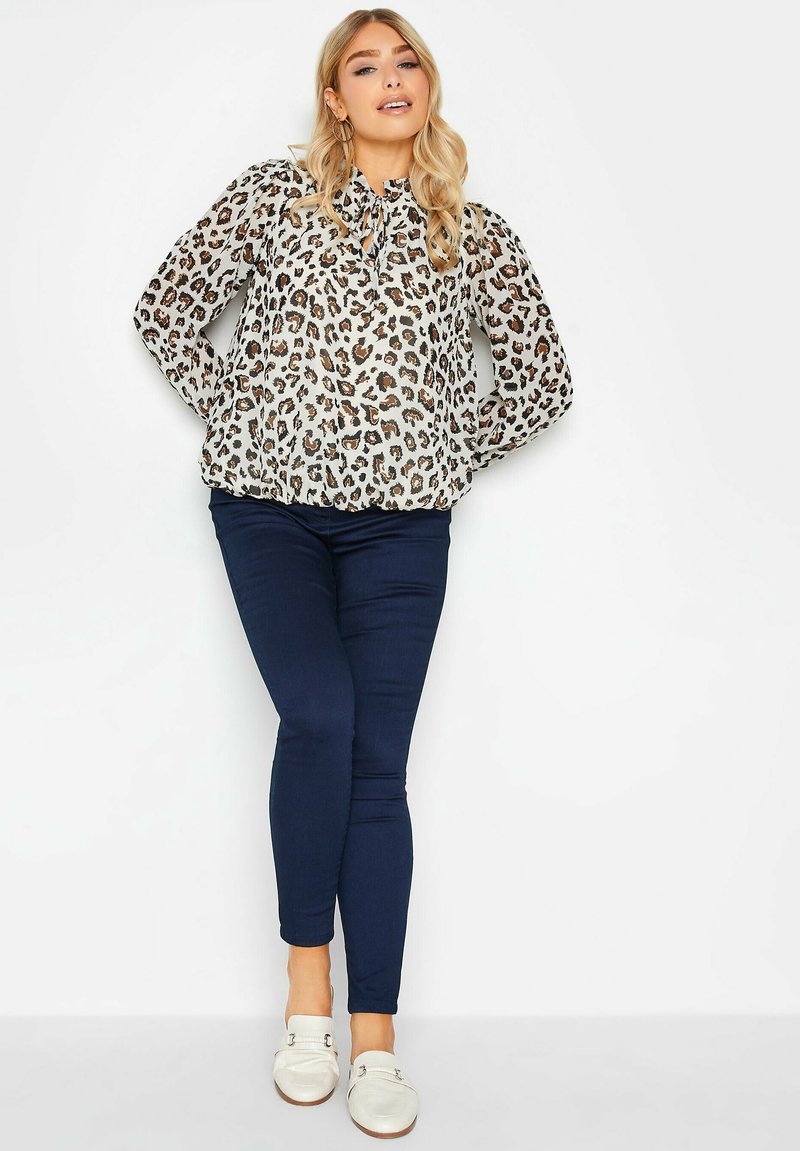 Blusa con stampa leopardata, collo a fiocco, materiale trasparente, maniche lunghe e puff; abbinata a jeans skinny blu scuro e scarpe slip-on bianche.