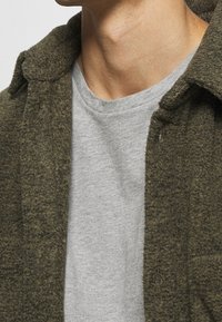 Olivově zelená, strukturovaná vlna košile s bundou a fleecovým povrchem, s límcem a knoflíkovým zapínáním, nošena přes světle šedé bavlněné tričko.