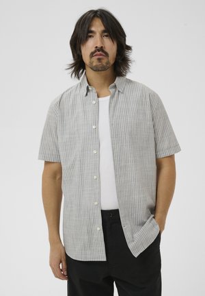 Mand med skulderlangt mørkt hår iført en lysegrå kortærmet skjorte med knapper over en hvid tanktop og sorte bukser.