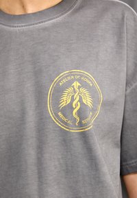 Camiseta de algodón gris con un logo circular amarillo que presenta un diseño de caduceo y palmera, acompañado del texto "ATELIER DE ROUPA SERVICIO MÉDICO."