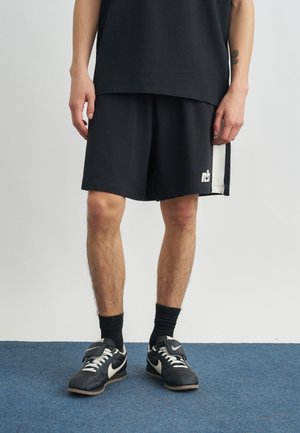 Mand iført sorte sportsshorts med hvide sidepaneler, sorte sokker og sorte Nike-sneakers, der står på blåt tæppe.
