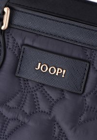 Zwart gewatteerd stof met een reliëfdesign, voorzien van een rechthoekig etiket met "JOOP!" in gouden letters. Gladde textuur met gedetailleerde stiksels.