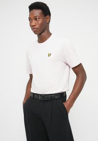 Hellrosa Baumwoll-T-Shirt mit kurzen Ärmeln, das ein kleines gelbes gesticktes Logo auf der linken Brust zeigt, kombiniert mit schwarzer Hose und Gürtel.