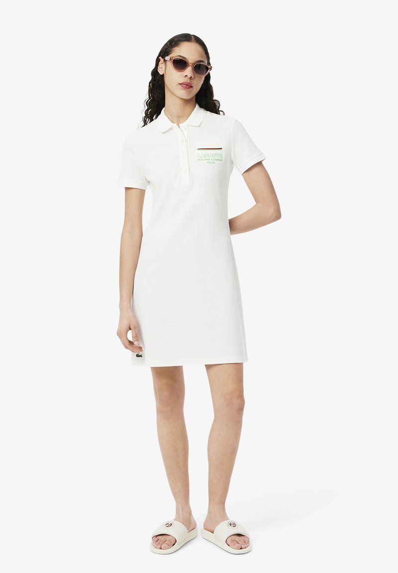 White polo dress with a collar and short sleeves, featuring a green logo design on the chest. Worn with white slide sandals.
