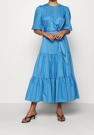 Femme portant une robe midi bleue à étages avec des manches bouffantes et une taille nouée, associée à des sandales noires à talons et un collier chaîne en or.