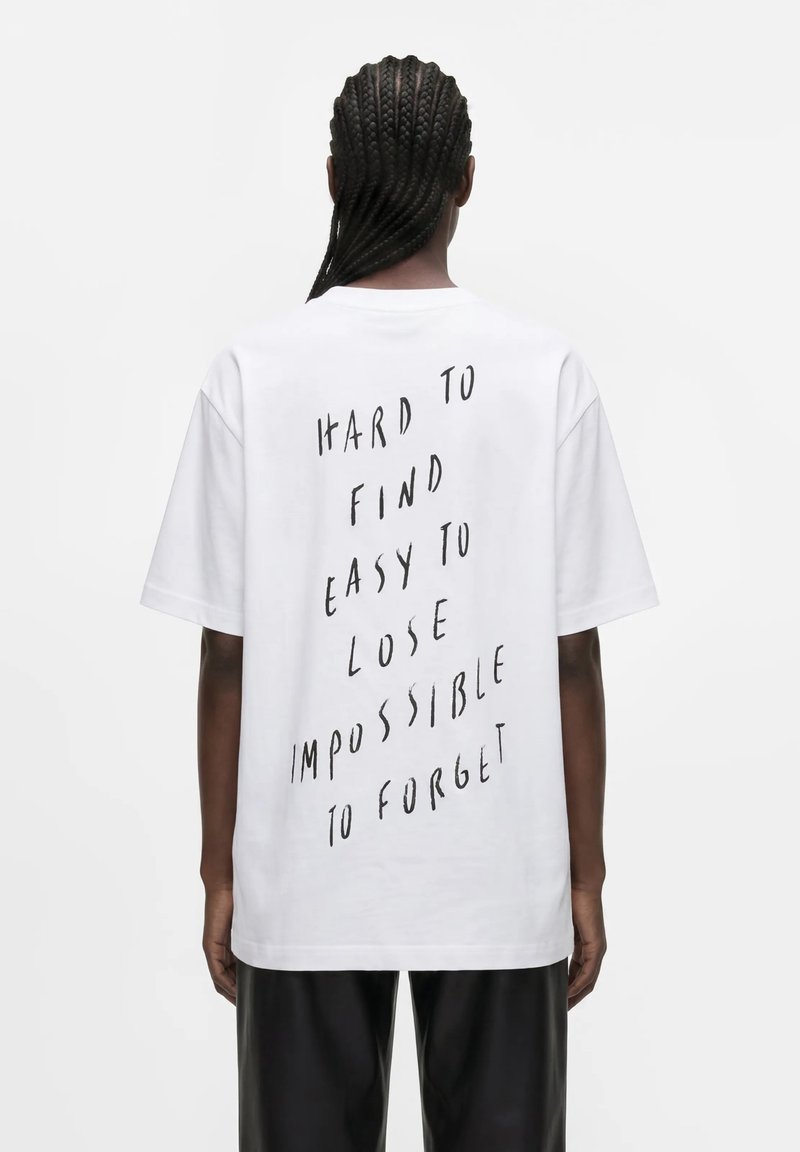 Άτομο με κοτσίδες που φοράει λευκό T-shirt με μαύρο κάθετο κείμενο που γράφει "Hard to find, easy to lose, impossible to forget" στην πλάτη.