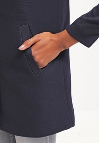 Marinblå kappa i strukturerad tyg, med en sidficka och avslappnad passform. Ärmen är trekvarts lång.