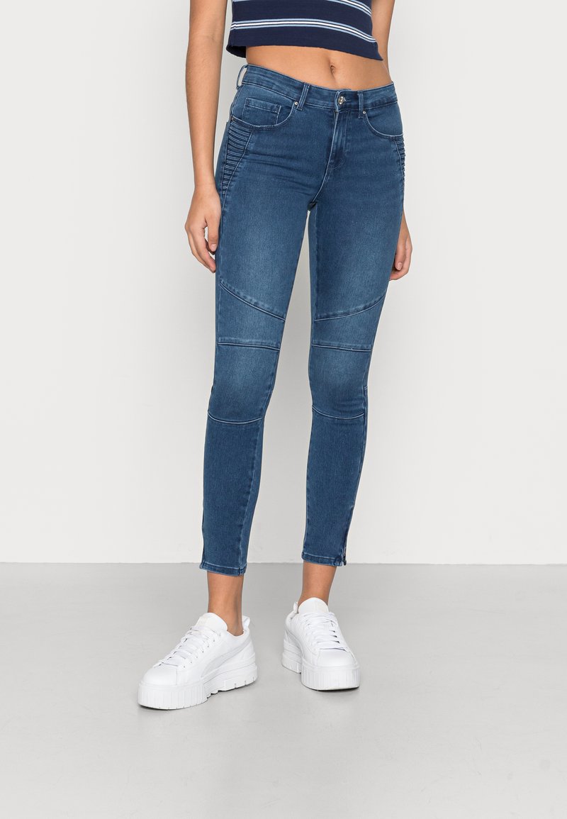 Jeans skinny em denim azul escuro com detalhes em painel e costura lateral. Comprimento até aos tornozelos, combinados com ténis de plataforma brancos.