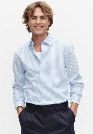Smilende ung mand med bølget blondt hår iført lyseblå stribet skjorte med knapper og mørke bukser, hænder i lommen, neutralt baggrund.