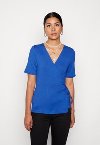 Top azul em tecido suave, com mangas curtas, decote em V e um detalhe de laço lateral para uma silhueta ajustada.