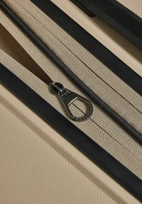Gros plan sur une surface en tissu beige texturé avec une fermeture éclair partiellement ouverte, dotée d'un tirette en métal argenté et de bords en caoutchouc noir.