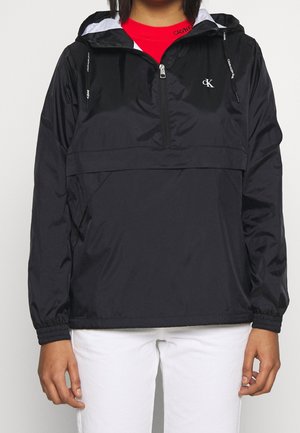 Veste noire à capuche avec fermeture éclair à l'avant et logos Calvin Klein portée sur un haut rouge et un pantalon blanc, montrée des épaules aux hanches.