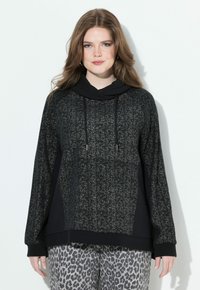 Joven con cabello ondulado, vestida con una sudadera negra moteada y pantalones gris con estampado de leopardo, de pie contra un fondo blanco liso.