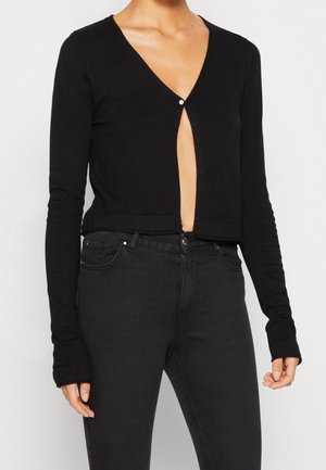 Frau trägt eine schwarze, kurz geschnittene Strickjacke mit einem oben befestigten Knopf und hoch taillierte schwarze Jeans.