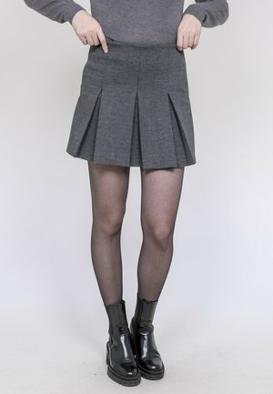 Personne portant une jupe plissée grise, des collants noirs transparents et des bottines noires, pointant vers le bas au niveau de la taille de la jupe.