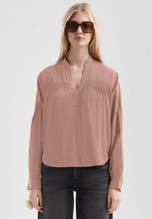 Femme aux longs cheveux blonds portant de grandes lunettes de soleil marron, un chemisier rose ample et un jean foncé, debout devant un fond clair uni.