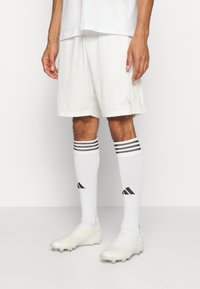 Pantalones cortos deportivos blancos con un pequeño logo beige, combinados con calcetas blancas hasta la rodilla que tienen rayas negras y logos negros, y zapatillas blancas.
