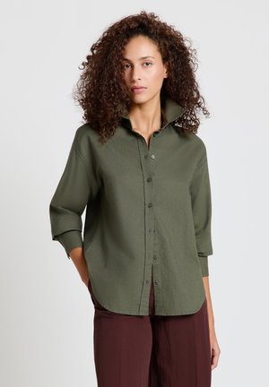 Donna con capelli ricci che indossa una camicia verde oliva con bottoni e maniche arrotolate e pantaloni marrone scuro su uno sfondo chiaro.