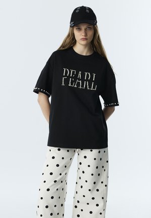Joven mujer con gorra negra, camiseta negra holgada con texto en perlas "DEAD PEARL" y pantalones blancos de pierna ancha con lunares negros.