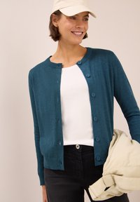 Teal gebreide cardigan met een ronde hals en zes knopen, gedragen over een wit shirt, gecombineerd met zwarte broek en een lichtgekleurde tas.