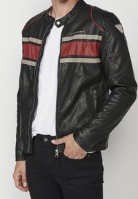 Uomo che indossa una giacca di pelle nera con strisce orizzontali rosse e bianche, camicia bianca e pantaloni neri, mano in tasca.