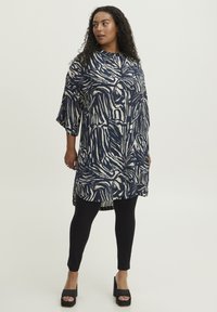 Tunica astratta a righe blu navy e crema con orlo asimmetrico, vestibilità comoda e maniche a 3/4, abbinata a leggings neri e scarpe con plateau.